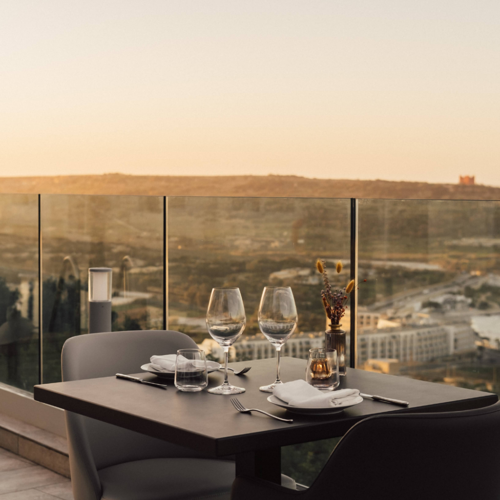A set up table with 2 wine glasses overlooking a view of Mellieha BAY