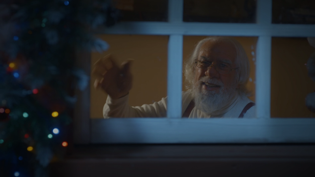 Santa Clause waving from a window which is decorated with Christmas lights