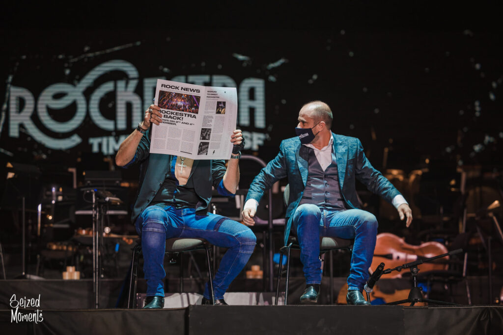 Two men sitting on chairs onstage. One is reading a newspaper, the other is looking at him
