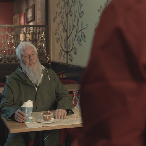 An old man with white hair and a white beard is smiling holding a coffee with foam and a muffin and sitting down at costa coffee